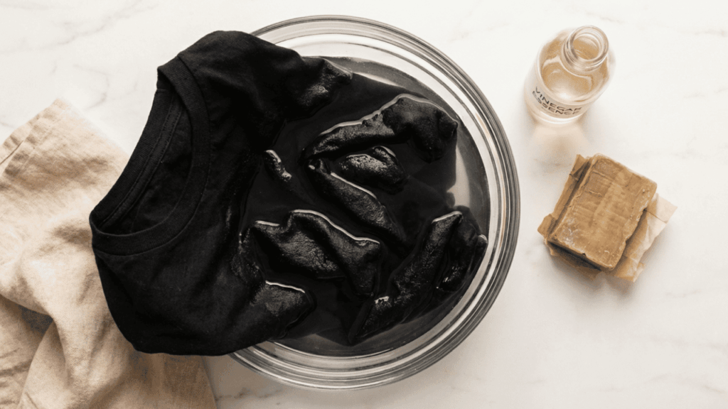 Schwarzes T-Shirt eingeweicht in einer Glasschüssel mit Wasser neben einer Flasche Essig und Gallseife – Hausmittel gegen Schweißflecken auf dunkler Kleidung